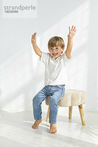 Happy boy with arms raised sitting on stool at home