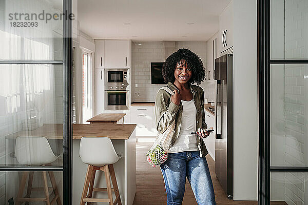 Smiling woman with mesh bag and smart phone standing at home