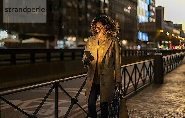 Lächelnde Frau hält in der Abenddämmerung Einkaufstüten mit Smartphone auf dem Fußweg