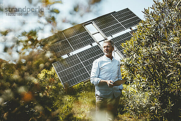 Reifer Mann steht an einem sonnigen Tag mit einem Tablet-Computer vor Sonnenkollektoren