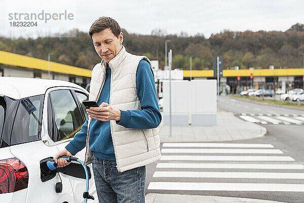 Mann benutzt Smartphone und steht neben Elektroauto am Bahnhof