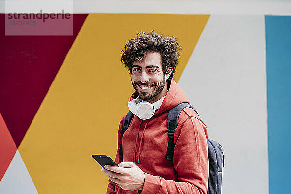 Lächelnder junger Mann mit Smartphone vor bunter Wand