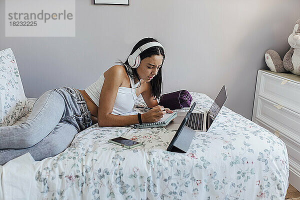 Mädchen mit Kopfhörern schreibt Notizen  liegt mit Gadgets zu Hause auf dem Bett