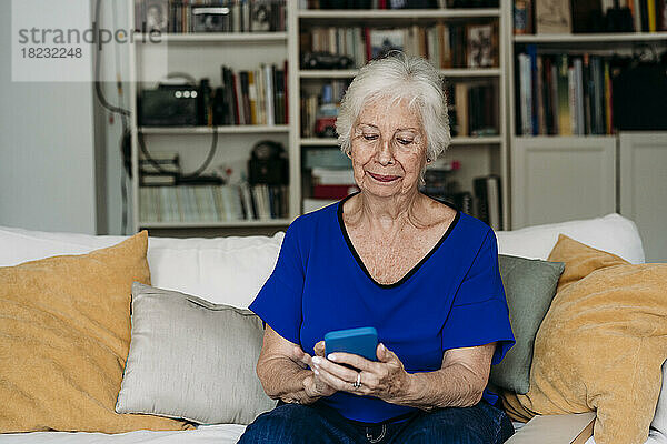 Lächelnde Seniorin benutzt Smartphone auf Sofa im Wohnzimmer