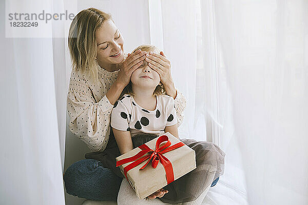 Mutter bedeckt Augen ihrer Tochter mit Weihnachtsgeschenk vor Vorhang
