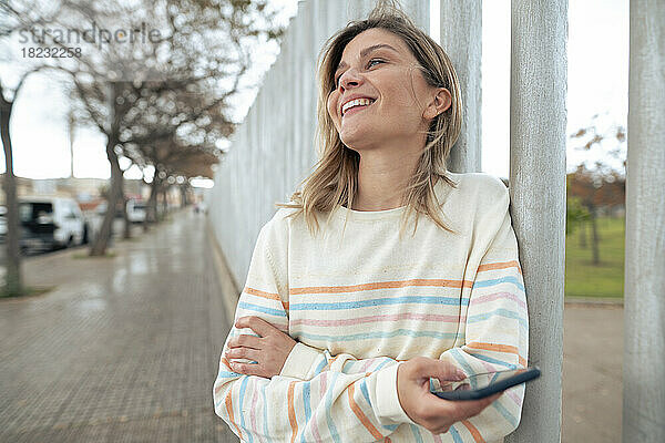 Glückliche blonde Frau hält ihr Smartphone am Zaun