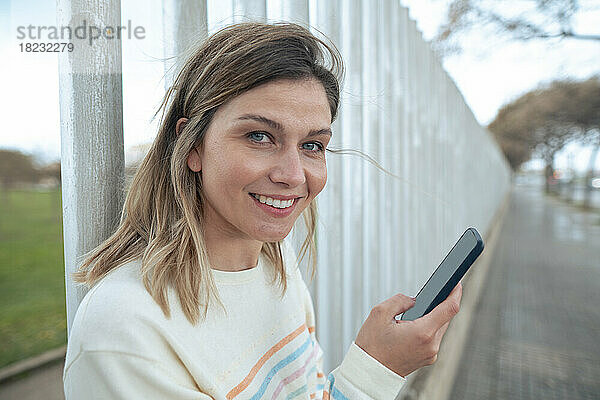 Lächelnde blonde Frau hält Smartphone in der Hand und lehnt am Zaun