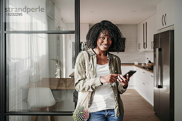 Junge Frau mit Smartphone und Netztasche steht zu Hause