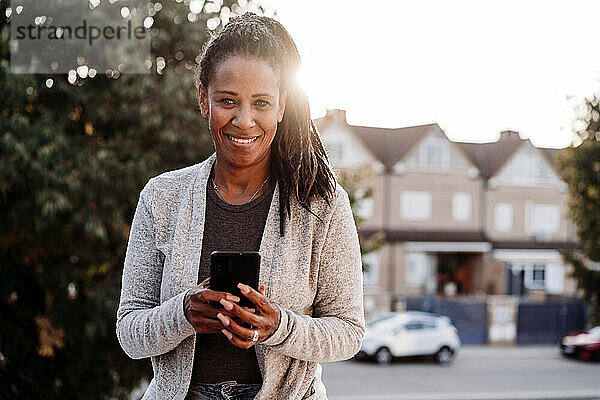 Glückliche Frau mit Smartphone steht bei Sonnenuntergang auf der Terrasse