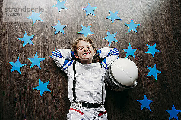 Glückliches süßes Mädchen im Weltraumkostüm  das zu Hause neben Helm und blauen Sternen auf dem Boden liegt