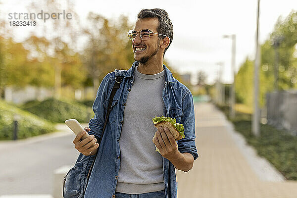 Fröhlicher junger Mann mit Sandwich und Smartphone steht auf Fußweg