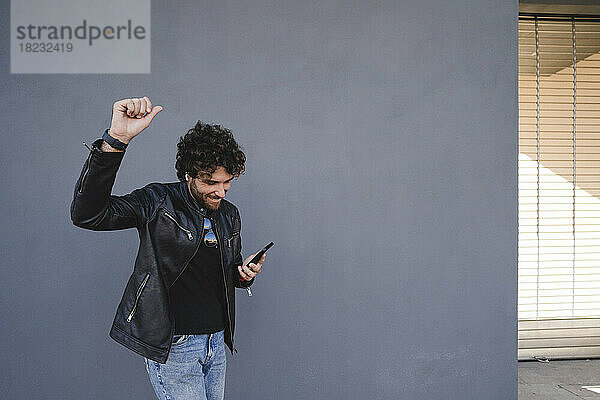 Glücklicher Mann mit erhobener Hand  der sein Mobiltelefon in der Hand hält und vor einer grauen Wand genießt