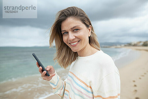 Lächelnde schöne blonde Frau  die am Strand ihr Smartphone benutzt