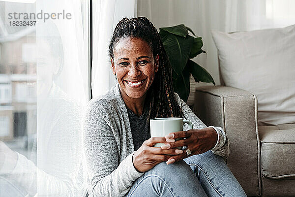Glückliche reife Frau  die zu Hause mit einer Kaffeetasse am Fenster sitzt