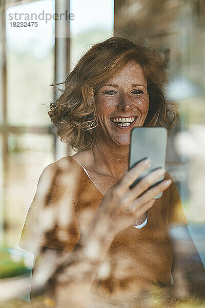 Glückliche Frau  die ihr Mobiltelefon benutzt und durch ein Glasfenster gesehen wird