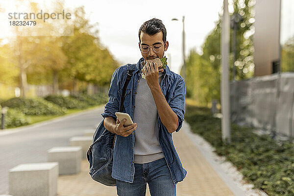 Junger Mann isst Sandwich und benutzt Smartphone auf Fußweg