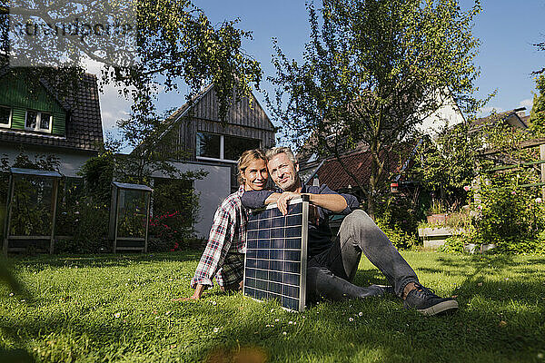 Lächelndes Paar sitzt mit Solarpanel auf Gras im Hinterhof