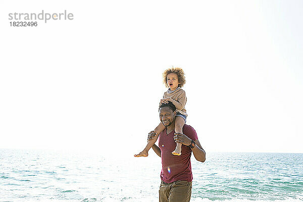 Glücklicher Mann mit Tochter auf Schultern  der am Strand vor dem Meer steht