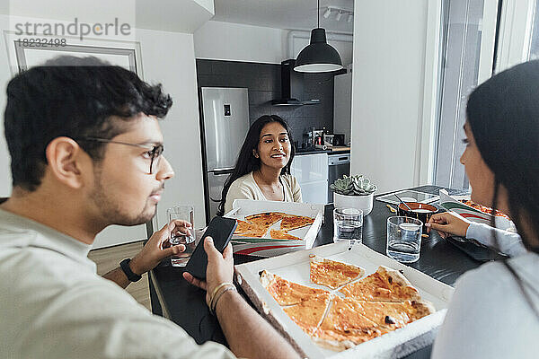 Mann mit Mobiltelefon sitzt mit Frauen zusammen und isst zu Hause Pizza