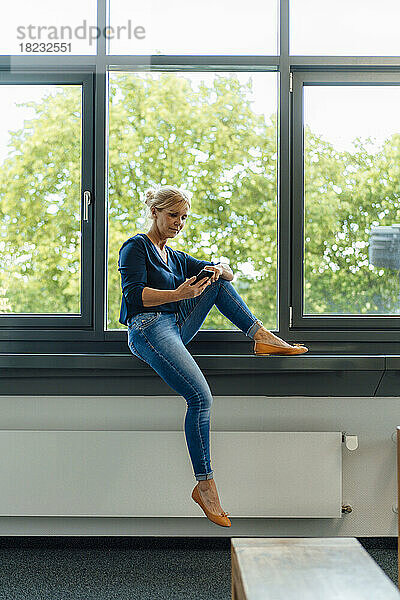 Geschäftsfrau sitzt mit Smartphone auf der Fensterbank im Büro
