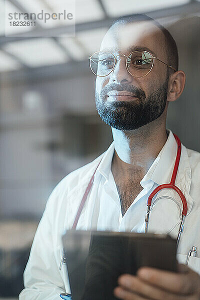 Smiling doctor holding tablet PC in hospital seen through glass