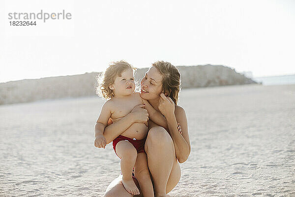 Lächelnde Mutter umarmt Sohn  der am Strand kniet und den Urlaub genießt