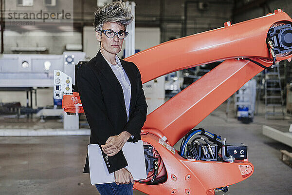 Selbstbewusste reife Geschäftsfrau mit Laptop steht neben Roboterarm in der Industrie