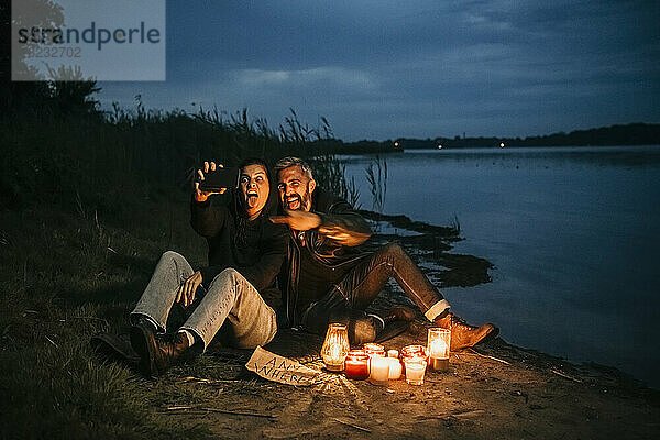 Glückliche Frau macht ein Selfie mit einem Mann  der bei brennenden Kerzen am Seeufer sitzt