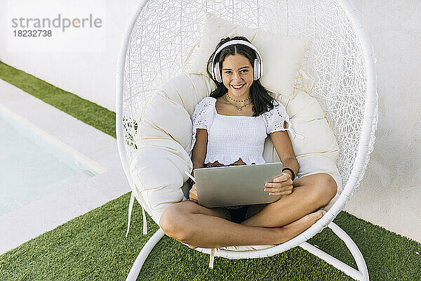 Fröhliches Mädchen mit Kopfhörern sitzt mit Laptop auf Hängesessel