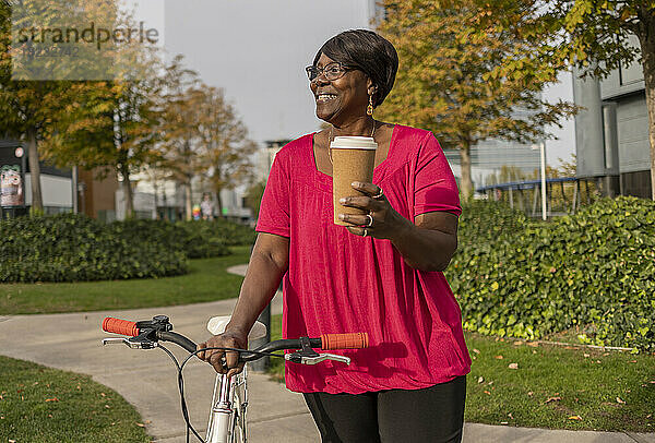 Lächelnde Frau mit Einwegbecher und Fahrrad im Park