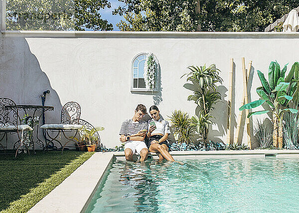 Teenager und Schwester sitzen an einem sonnigen Tag am Pool und nutzen ihr Smartphone