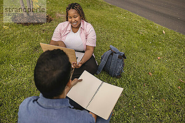 Happy young couple with laptop and book sitting on grass