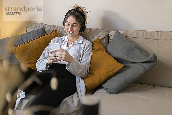 Glückliche junge Frau mit kabellosen Kopfhörern und Smartphone auf dem heimischen Sofa
