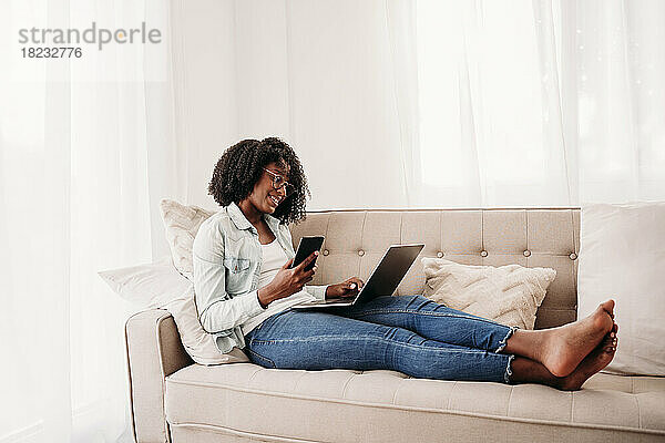 Junger Freiberufler mit Smartphone und Laptop sitzt zu Hause auf dem Sofa