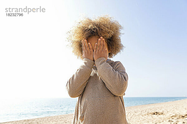 Mädchen bedeckt Gesicht mit Händen und steht vor dem Meer am Strand