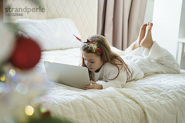 Mädchen mit Weihnachtsstirnband liegt auf dem Bett und benutzt zu Hause Laptop
