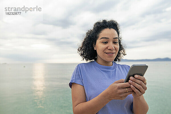 Lächelnde Frau  die ihr Mobiltelefon vor dem Meer benutzt