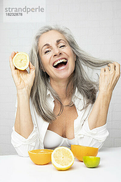 Happy mature woman sitting with oranges and lemons at table in front of wall