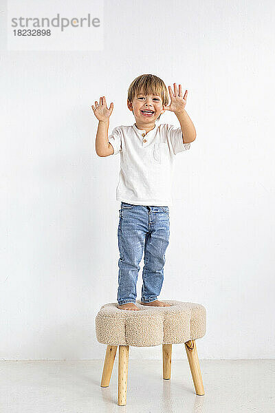 Smiling boy gesturing on stool in front of wall at home