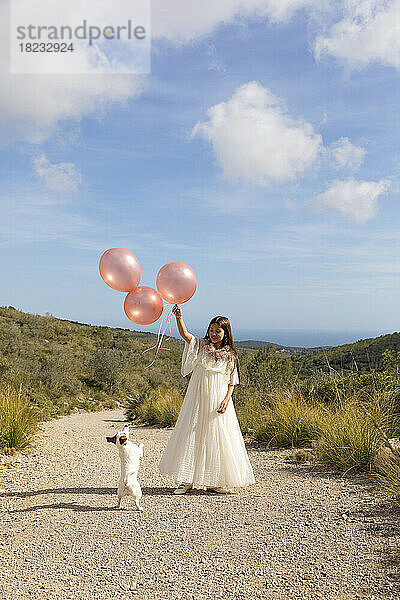 Mädchen in weißem Kleid hält Luftballons und spielt mit Hund auf dem Weg