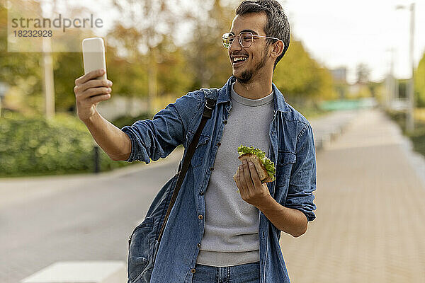 Glücklicher junger Mann  der am Fußweg ein Selfie mit dem Smartphone macht
