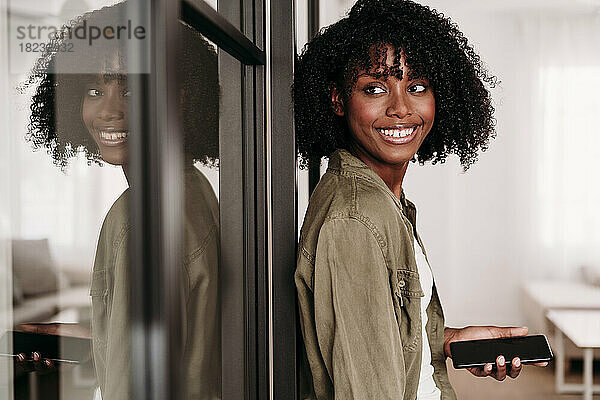 Lächelnde Frau mit Smartphone lehnt zu Hause an Glastür