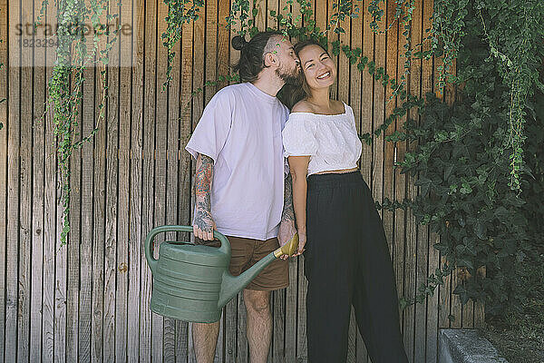 Hipster-Mann hält Gießkanne in der Hand und küsst glückliche Freundin vor Holzwand