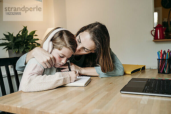 Mutter küsst Tochter  die zu Hause am Tisch Hausaufgaben macht