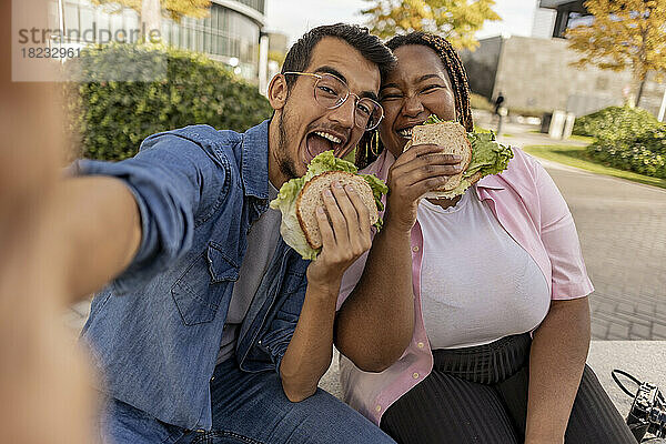 Glückliches junges Paar  das Sandwich isst und ein Selfie auf dem Fußweg macht