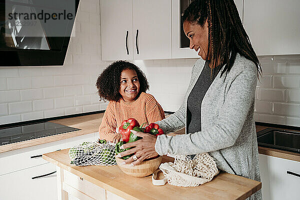 Lächelndes Mädchen mit Mutter  die zu Hause Gemüse auf dem Tisch hält