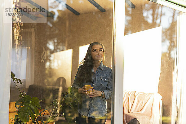 Reife Frau mit Kaffeetasse und Blick aus dem Fenster eines Hauses