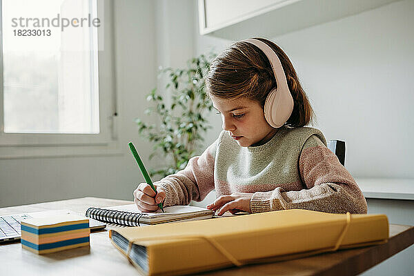 Mädchen mit kabellosen Kopfhörern macht Hausaufgaben und sitzt zu Hause am Tisch