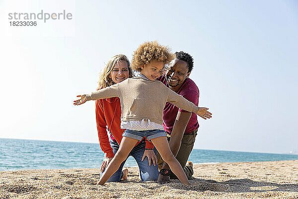 Glückliches Mädchen  das mit ausgestreckten Armen vor den Eltern am Strand steht