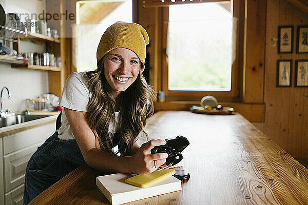 Glückliche junge Frau mit Kamera lehnt an der Küchentheke